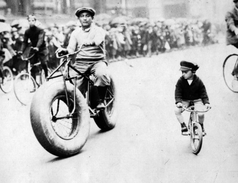 Una persona pedalea subido a una fat bike, mientras un niño le mira. Foto en blanco y negro, del siglo XIX.